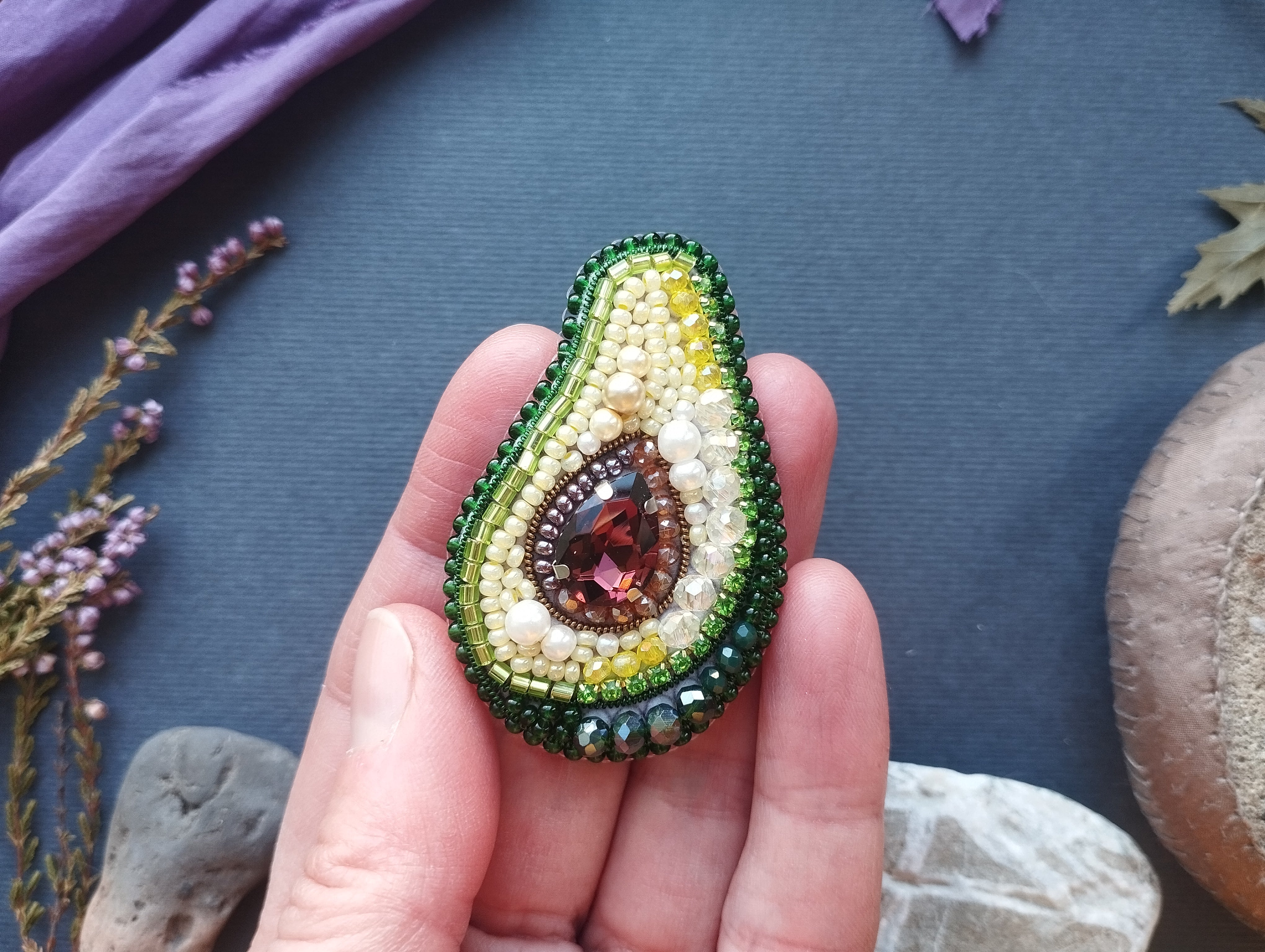 A hand holding a beaded avocado-shaped brooch with a gemstone in the center, surrounded by green and yellow beads.